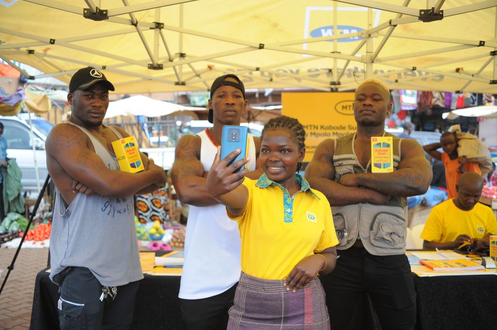 A lady poses for a selfie with the MTN Kabode Supa Smartphone standing next to Kabode men. Kabode stands for strong meaning the phone is built to last long